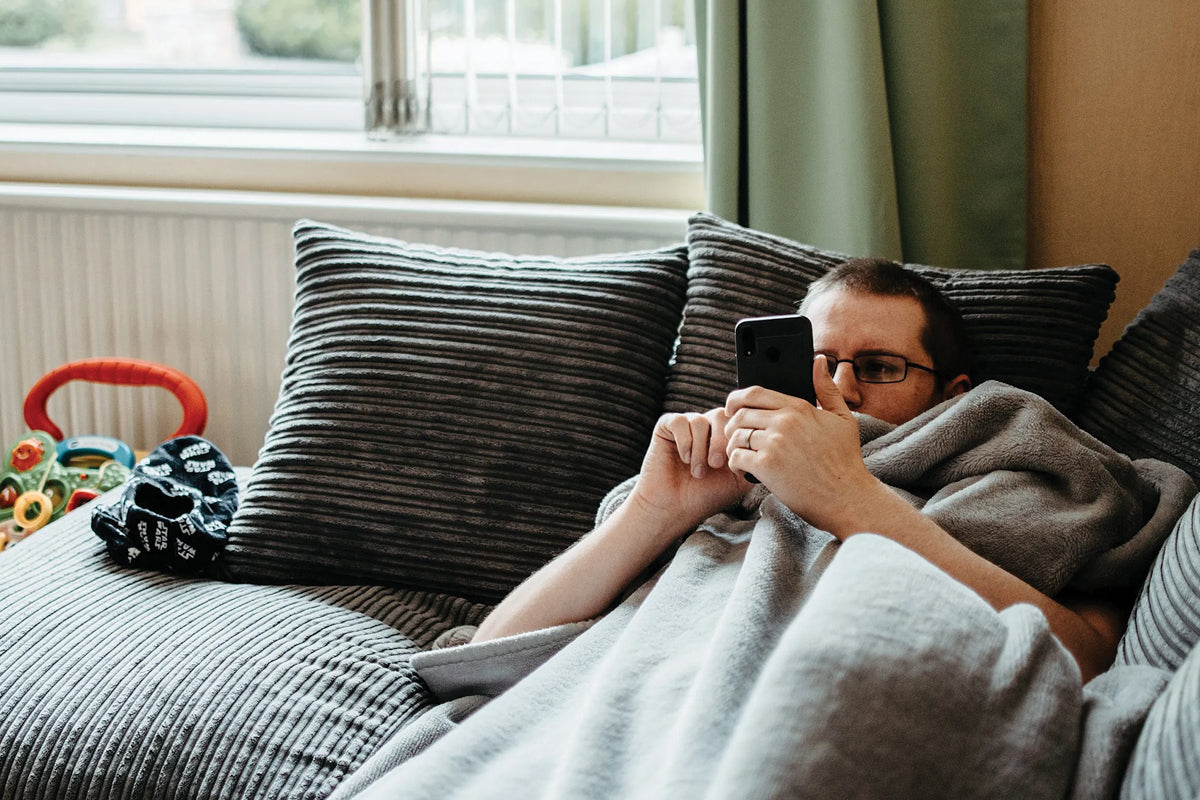 A man lying on the couch, scrolling through his phone and  looking lethargic.