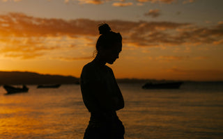 A woman looking down, finding mental calmness by the lake at sunset, surrounded by nature’s serenity.