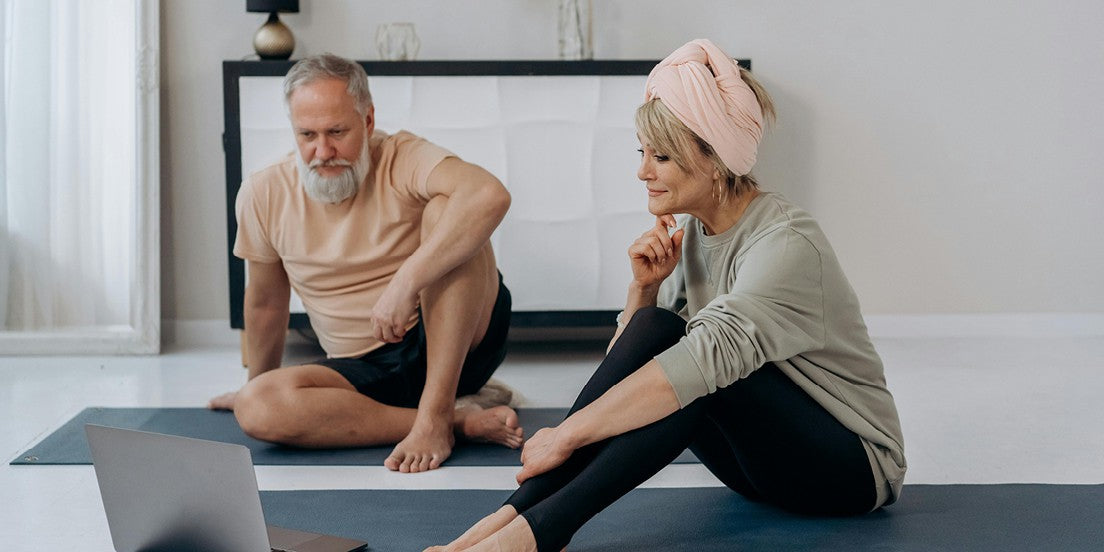 A man and a woman sitting on yoga mats, following an online exercise class for overall body health, wellness, and vitality.